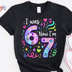 May include: Black t-shirt featuring the text "I was 6 Now I'm 7" in rainbow colours. The design includes hearts, stars, and birthday cake graphics. White trainers are visible in the bottom right corner.