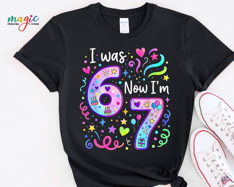 May include: Black t-shirt featuring the text "I was 6 Now I'm 7" in rainbow colours. The design includes hearts, stars, and birthday cake graphics. White trainers are visible in the bottom right corner.