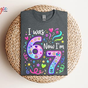 May include: Dark grey t-shirt featuring the text "I was 6 Now I'm 7" in a vibrant, playful design. The graphic includes hearts, stars, balloons, and cake illustrations, ideal for a seventh birthday celebration.