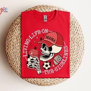 May include: Red t-shirt featuring a white graphic of a skeleton wearing a red baseball cap that says "Mama" and holding a coffee cup. The graphic also includes a football and a football goal. The text on the shirt says "Living life on the sidelines".