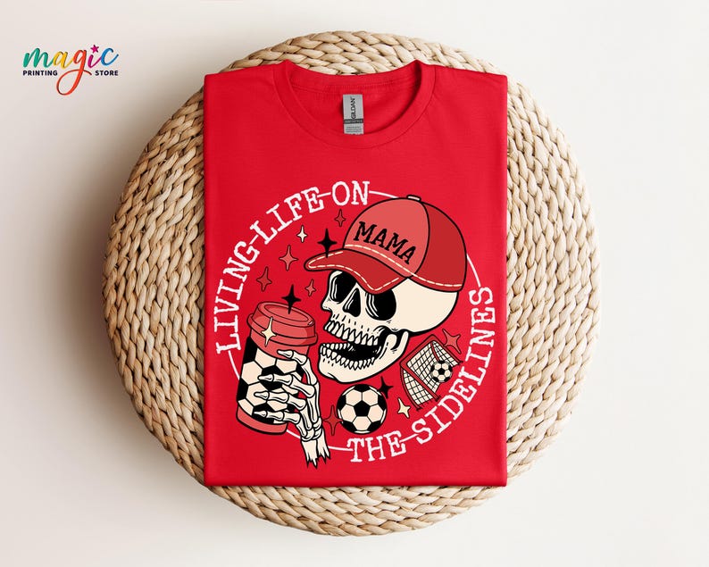 May include: Red t-shirt featuring a white graphic of a skeleton wearing a red baseball cap that says "Mama" and holding a coffee cup. The graphic also includes a football and a football goal. The text on the shirt says "Living life on the sidelines".