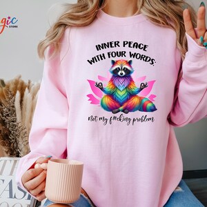 Pu&ograve; includere: Felpa rosa chiaro con la scritta "INNER PEACE WITH FOUR WORDS: Not my f#cking problem." Un procione colorato in posizione del loto &egrave; al centro. La felpa ha le maniche lunghe.
