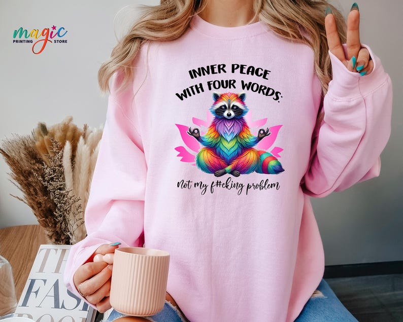 Pu&ograve; includere: Felpa rosa chiaro con la scritta "INNER PEACE WITH FOUR WORDS: Not my f#cking problem." Un procione colorato in posizione del loto &egrave; al centro. La felpa ha le maniche lunghe.