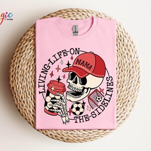 May include: Pink t-shirt featuring a graphic of a skull wearing a red baseball cap that says "Mama" and holding a coffee cup. The skull is holding a football and the text "Living life on the sidelines" is around the skull.