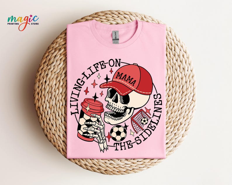May include: Pink t-shirt featuring a graphic of a skull wearing a red baseball cap that says "Mama" and holding a coffee cup. The skull is holding a football and the text "Living life on the sidelines" is around the skull.
