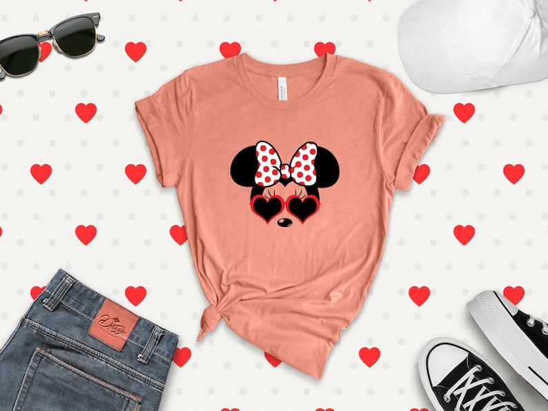 May include: A pink t-shirt with a graphic of Minnie Mouse wearing heart-shaped sunglasses and a red and white polka dot bow. The background is a white pattern of red hearts.