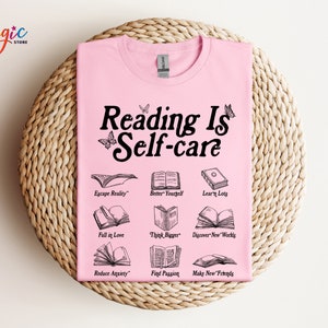 May include: Pink t-shirt with the text "Reading Is Self-care" in black. The shirt features illustrations of open books with text describing the benefits of reading, such as "Escape Reality", "Better Yourself", "Learn Lots", "Fall in Love", "Think Bigger", "Discover New Worlds", "Reduce Anxiety", "Find Passion", and "Make New Friends".