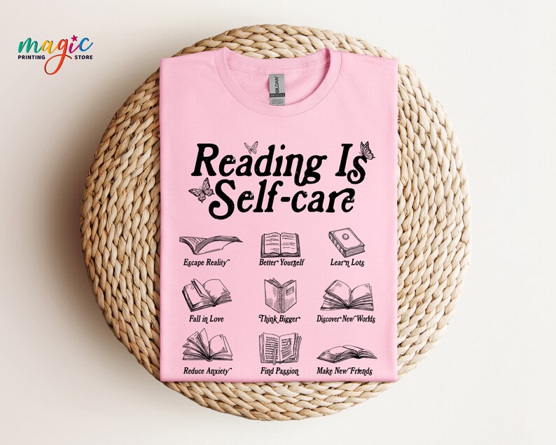May include: Pink t-shirt with the text "Reading Is Self-care" in black. The shirt features illustrations of open books with text describing the benefits of reading, such as "Escape Reality", "Better Yourself", "Learn Lots", "Fall in Love", "Think Bigger", "Discover New Worlds", "Reduce Anxiety", "Find Passion", and "Make New Friends".