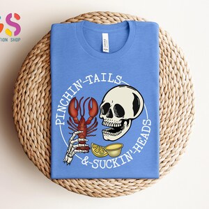K&ouml;nnte beinhalten: Ein hellblaues T-Shirt mit einem wei&szlig;en Grafikdesign, das einen Sch&auml;del mit einer Hummerklaue im Maul, Zitronen und den Text "Pinchin' Tails & Suckin' Heads" zeigt.