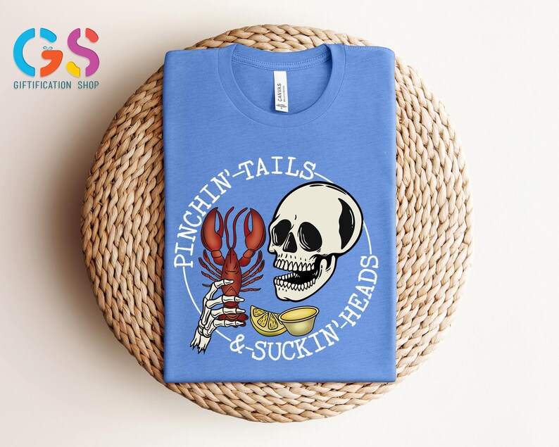 K&ouml;nnte beinhalten: Ein hellblaues T-Shirt mit einem wei&szlig;en Grafikdesign, das einen Sch&auml;del mit einer Hummerklaue im Maul, Zitronen und den Text "Pinchin' Tails & Suckin' Heads" zeigt.
