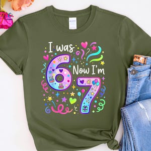 May include: Khaki green t-shirt featuring the phrase "I was 6 Now I'm 7" in a vibrant, celebratory design. The graphic includes the numbers 6 and 7, adorned with stars, hearts, balloons, and confetti, ideal for a birthday party.