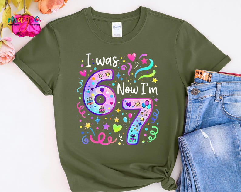 May include: Khaki green t-shirt featuring the phrase "I was 6 Now I'm 7" in a vibrant, celebratory design. The graphic includes the numbers 6 and 7, adorned with stars, hearts, balloons, and confetti, ideal for a birthday party.