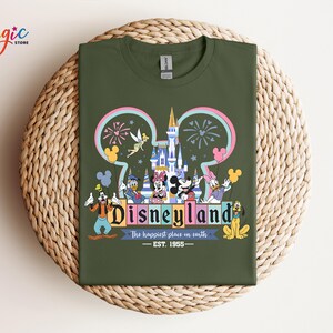 Puede incluir: Una camiseta verde oliva con un gr&aacute;fico colorido de Disneyland. El dise&ntilde;o incluye el logotipo de Disneyland, personajes, un castillo, fuegos artificiales y el texto "The happiest place on earth - EST. 1955."