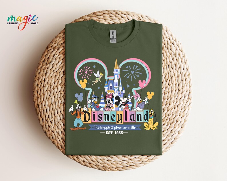 Puede incluir: Una camiseta verde oliva con un gr&aacute;fico colorido de Disneyland. El dise&ntilde;o incluye el logotipo de Disneyland, personajes, un castillo, fuegos artificiales y el texto "The happiest place on earth - EST. 1955."
