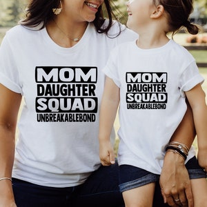 May include: White t-shirts with black text that reads "Mom Daughter Squad Unbreakablebond". The shirts are worn by a woman and a young girl.
