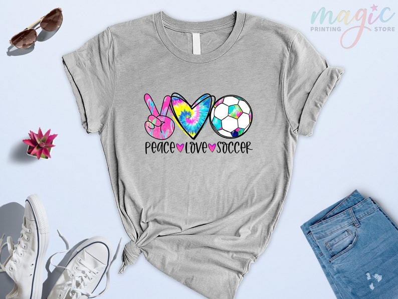 Może przedstawiać: Szara koszulka z nadrukiem znaku pokoju, serca i piłki nożnej. Nadruk wykonany jest w stylu tie-dye w kolorach r&oacute;żowym, niebieskim i ż&oacute;łtym. Pod nadrukiem znajduje się napis "Peace Love Soccer".