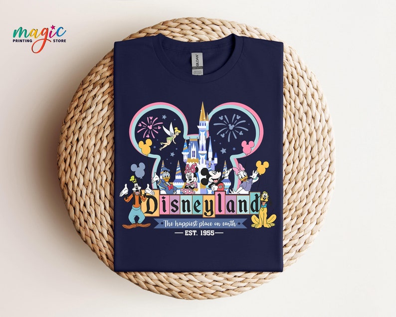 Puede incluir: Camiseta azul marino con un dise&ntilde;o colorido de Disneyland. El dise&ntilde;o incluye el logotipo de Disneyland, Mickey Mouse y el texto "The happiest place on earth - EST. 1955". La camiseta est&aacute; doblada sobre una superficie tejida.