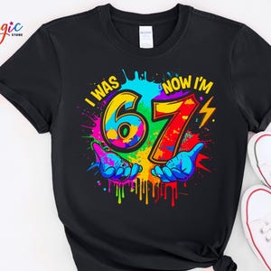 May include: Black t-shirt with a colorful graphic that reads "I WAS 67 NOW I'M" with the numbers 67 in a vibrant design. The design includes two blue hands and a lightning bolt. White sneakers are visible.