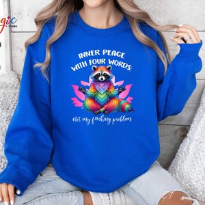 Pu&ograve; includere: Una felpa blu reale con un procione arcobaleno che medita su un fiore di loto. Il testo sulla felpa recita: "INNER PEACE WITH FOUR WORDS: Not my f*cking problem."