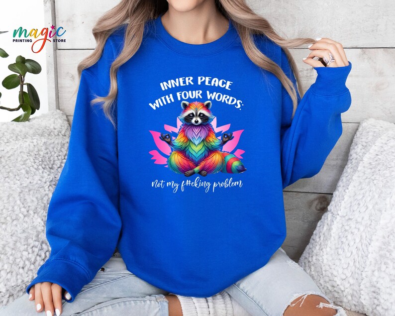 Pu&ograve; includere: Una felpa blu reale con un procione arcobaleno che medita su un fiore di loto. Il testo sulla felpa recita: "INNER PEACE WITH FOUR WORDS: Not my f*cking problem."
