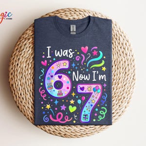May include: A navy blue t-shirt featuring the text "I was 6 Now I'm 7". The design incorporates colourful illustrations of hearts, stars, balloons, and cakes. The shirt is displayed on a woven, circular surface.