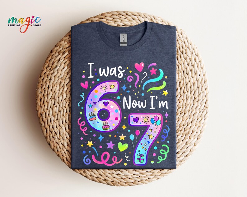 May include: A navy blue t-shirt featuring the text "I was 6 Now I'm 7". The design incorporates colourful illustrations of hearts, stars, balloons, and cakes. The shirt is displayed on a woven, circular surface.