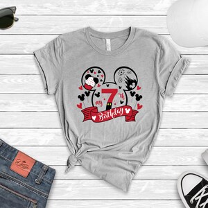 以下が含まれることがあります： 赤と黒のミッキーマウスのデザインが施されたグレーのTシャツ。デザインは、右の耳に数字の7が描かれたミッキーマウスの耳と、赤いリボンに「my 7th birthday」という文字が書かれています。