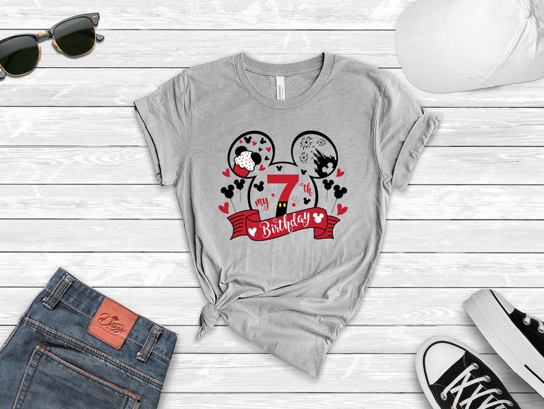 以下が含まれることがあります： 赤と黒のミッキーマウスのデザインが施されたグレーのTシャツ。デザインは、右の耳に数字の7が描かれたミッキーマウスの耳と、赤いリボンに「my 7th birthday」という文字が書かれています。