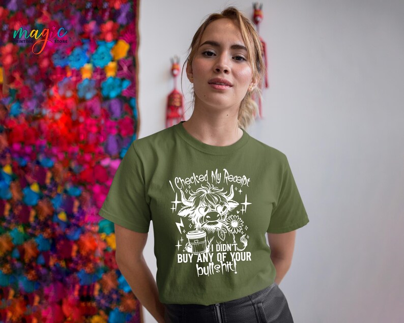 May include: Olive green t-shirt with a white graphic of a Highland cow, coffee cup, and flower. The text on the shirt reads "I checked my receipt, I didn't buy any of your bullshit!" The shirt is a casual style.