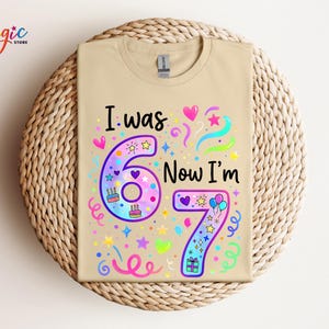 May include: A beige t-shirt featuring the phrase "I was 6, Now I'm 7" in a vibrant, celebratory design. The graphic includes hearts, stars, confetti, and cake illustrations, ideal for a child's seventh birthday.