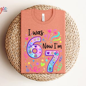 May include: A peach-coloured t-shirt with the text "I was 6 Now I'm 7" in a colourful design. The design includes the numbers 6 and 7, hearts, stars, balloons, and confetti. The shirt is displayed on a woven, circular mat.