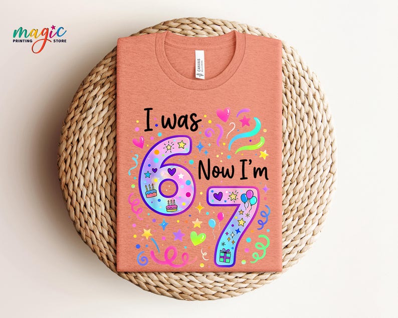 May include: A peach-coloured t-shirt with the text "I was 6 Now I'm 7" in a colourful design. The design includes the numbers 6 and 7, hearts, stars, balloons, and confetti. The shirt is displayed on a woven, circular mat.