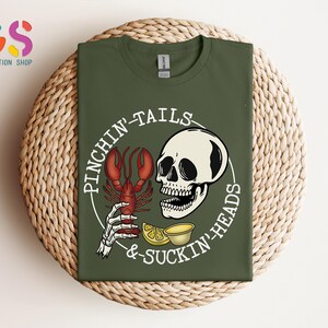 K&ouml;nnte beinhalten: Gr&uuml;nes T-Shirt mit einer wei&szlig;en Grafik eines Sch&auml;dels, der einen Hummer mit dem Text "Pinchin'-Tails &-Suckin'-Heads" h&auml;lt.