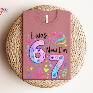 May include: A dusty rose t-shirt featuring the text "I was 6 Now I'm 7" in a vibrant design. The design incorporates the numbers 6 and 7, hearts, stars, balloons, and confetti. The t-shirt is presented on a woven, circular placemat.