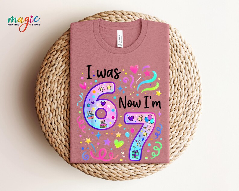 May include: A dusty rose t-shirt featuring the text "I was 6 Now I'm 7" in a vibrant design. The design incorporates the numbers 6 and 7, hearts, stars, balloons, and confetti. The t-shirt is presented on a woven, circular placemat.