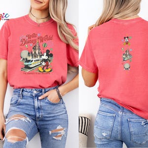 May include: Red t-shirt with a Walt Disney World graphic on the front, showing Mickey Mouse, a train, and a castle. The back has a smaller graphic with Mickey Mouse and Disney World icons. The front text reads "Walt Disney World" and "The Happiest Place on Earth."