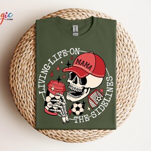 May include: Green t-shirt featuring a graphic of a skeleton wearing a red baseball cap that says "Mama" and holding a coffee cup. The graphic also includes a football and a football goal. The text on the shirt says "Living Life On The Sidelines".