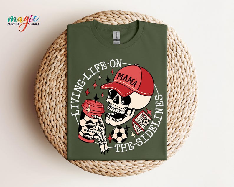 May include: Green t-shirt featuring a graphic of a skeleton wearing a red baseball cap that says "Mama" and holding a coffee cup. The graphic also includes a football and a football goal. The text on the shirt says "Living Life On The Sidelines".