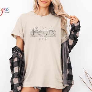 May include: Beige t-shirt with a desert landscape graphic featuring cacti, stars, and the text "If the stars were made to worship, so will I." The shirt is paired with a black and gray plaid shirt.