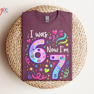 May include: A maroon t-shirt with the text "I was 6 Now I'm 7" in white. The numbers are decorated with colourful stars, hearts, and balloons. The shirt is displayed on a woven, circular mat.