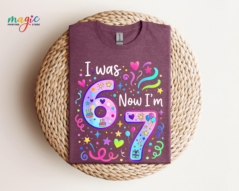 May include: A maroon t-shirt with the text "I was 6 Now I'm 7" in white. The numbers are decorated with colourful stars, hearts, and balloons. The shirt is displayed on a woven, circular mat.