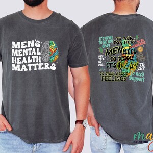 Może przedstawiać: Szary t-shirt z białym nadrukiem z przodu, kt&oacute;ry głosi "Men's Mental Health Matters" i kolorową grafiką m&oacute;zgu. Z tyłu t-shirtu znajduje się kolorowa grafika z tekstem "It's okay to ask for help. Men need to know it's okay to talk about feelings. To need support."