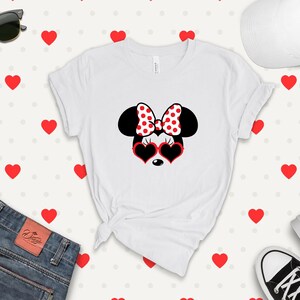 May include: White t-shirt with a black and white illustration of Minnie Mouse wearing red heart-shaped sunglasses and a red and white polka dot bow. The background is a white pattern of red hearts.