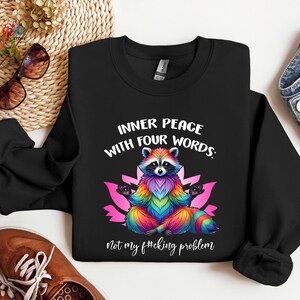 Pu&ograve; includere: Felpa nera con una grafica colorata di un procione in posa yoga. Il testo recita: "INNER PEACE WITH FOUR WORDS: Not my f#cking problem."