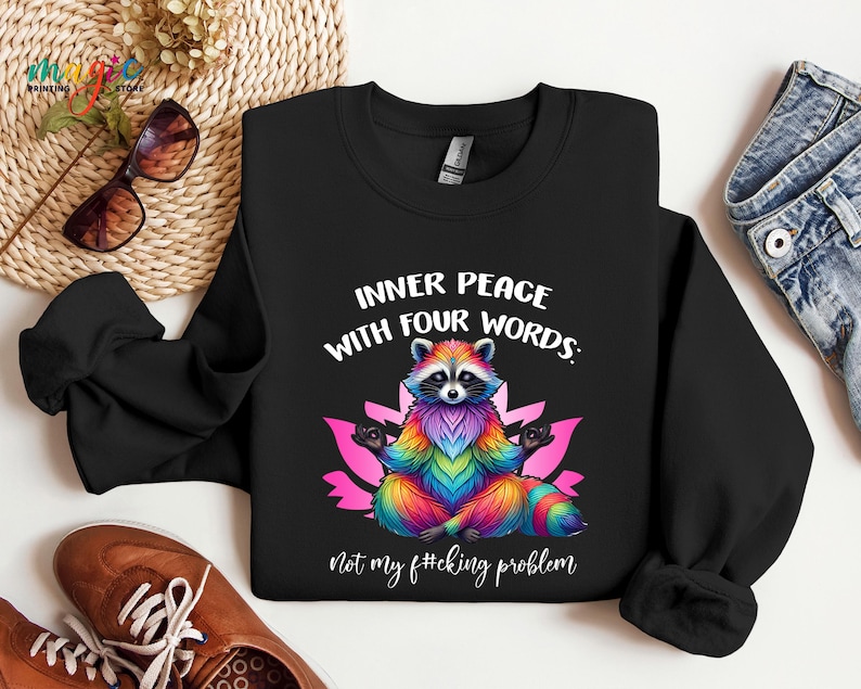 Pu&ograve; includere: Felpa nera con una grafica colorata di un procione in posa yoga. Il testo recita: "INNER PEACE WITH FOUR WORDS: Not my f#cking problem."