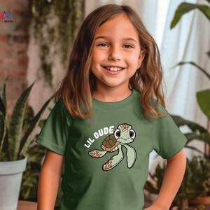 May include: A green t-shirt featuring a cartoon turtle with the text "Lil Dude". The turtle has a brown shell and green flippers. The shirt is made of a soft material.