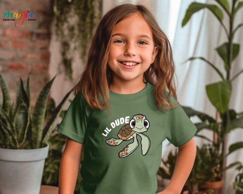 May include: A green t-shirt featuring a cartoon turtle with the text "Lil Dude". The turtle has a brown shell and green flippers. The shirt is made of a soft material.