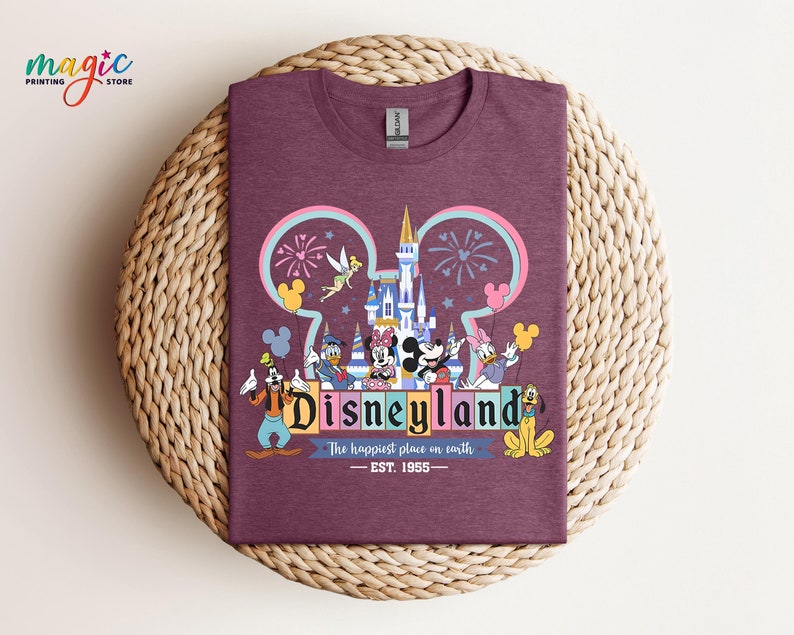 Puede incluir: Una camiseta granate con un gr&aacute;fico colorido de Disneyland. El dise&ntilde;o incluye el logotipo de Disneyland, un castillo, fuegos artificiales y varios personajes de Disney. El texto dice "Disneyland" y "The happiest place on earth - EST. 1955."