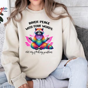 Pu&ograve; includere: Felpa beige con un procione colorato che medita su un fiore di loto rosa. Il testo recita: "INNER PEACE WITH FOUR WORDS: Not my f#cking problem."