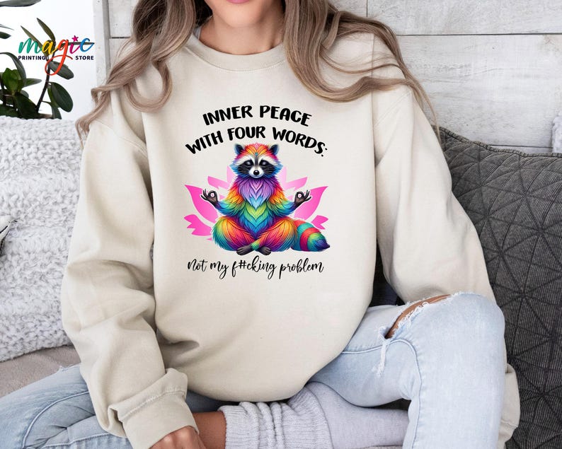 Pu&ograve; includere: Felpa beige con un procione colorato che medita su un fiore di loto rosa. Il testo recita: "INNER PEACE WITH FOUR WORDS: Not my f#cking problem."